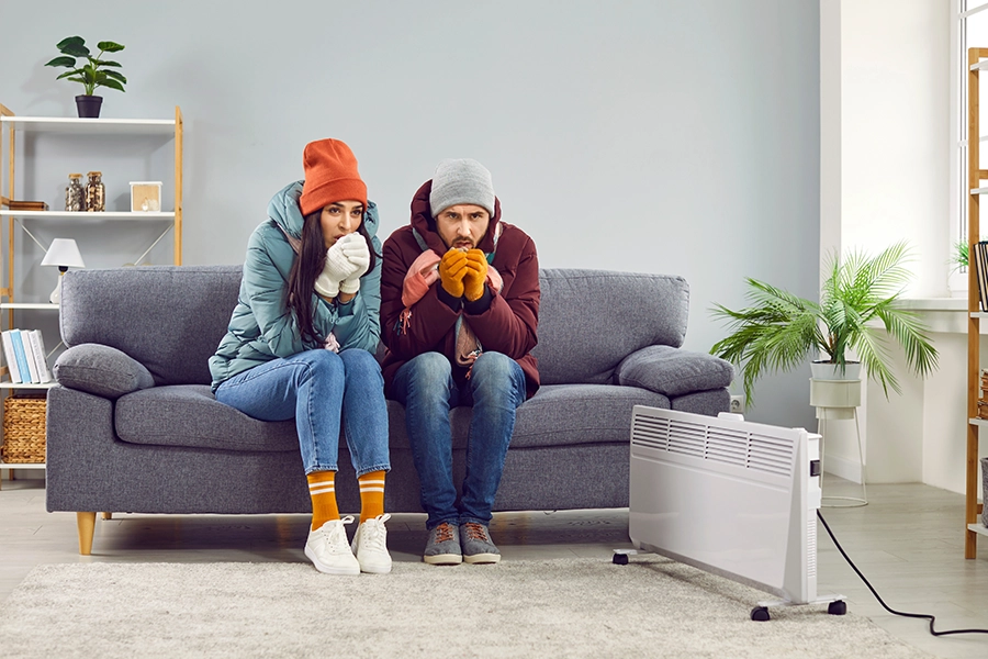 A young couple bundled up in their Lincoln, IL home in coats, hats, and gloves, huddled around their heater because their HVAC needs a professional heating repairA young couple bundled up in their Lincoln, IL home in coats, hats, and gloves, huddled around their heater because their HVAC needs a professional heating repair